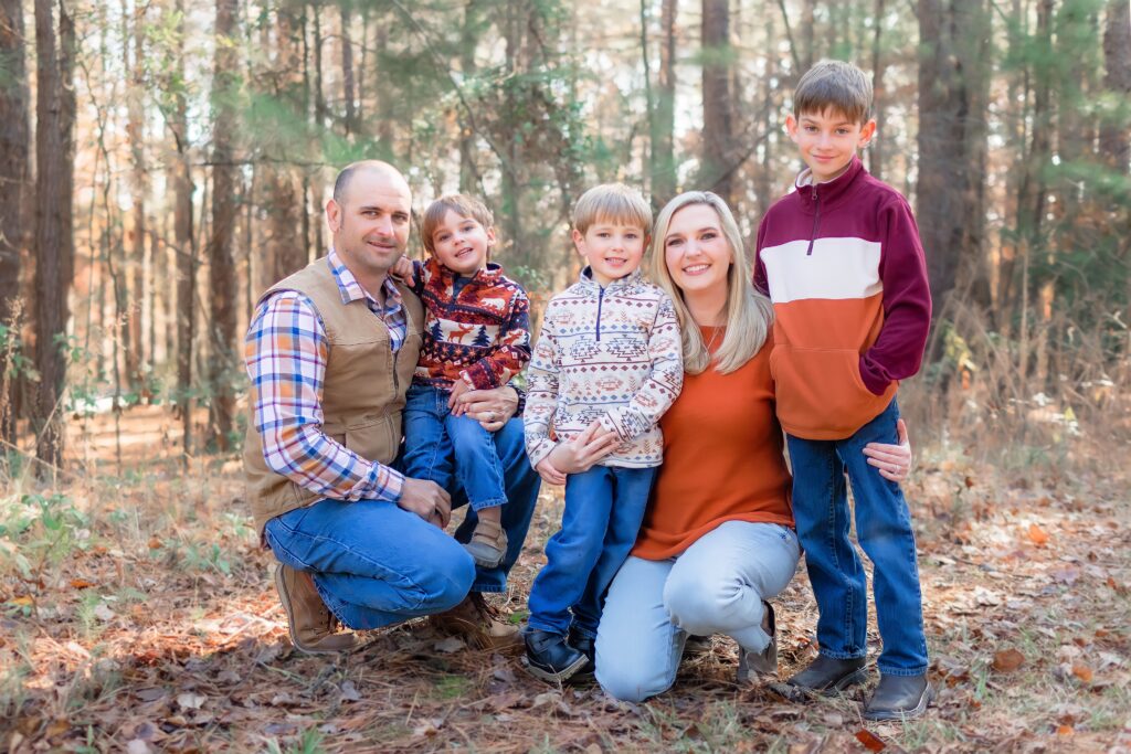 Family embracing and smiling in wooded clearing with trees in the background.