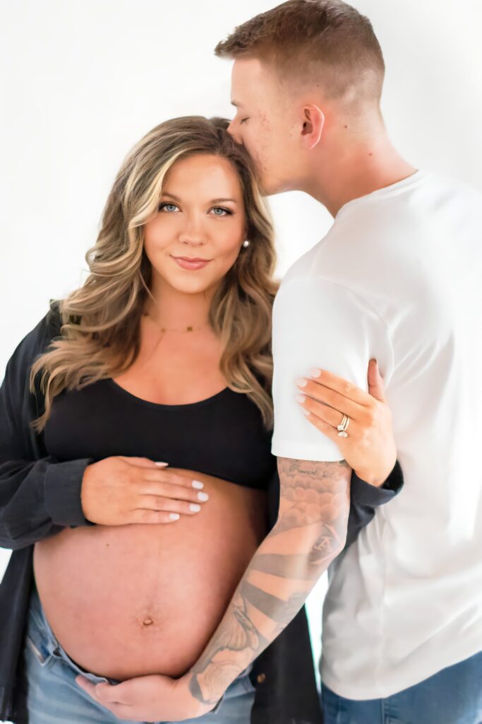 Maternity photo with husband kissing wife’s forehead while cradling her bare baby bump, filled with emotion and connection.
