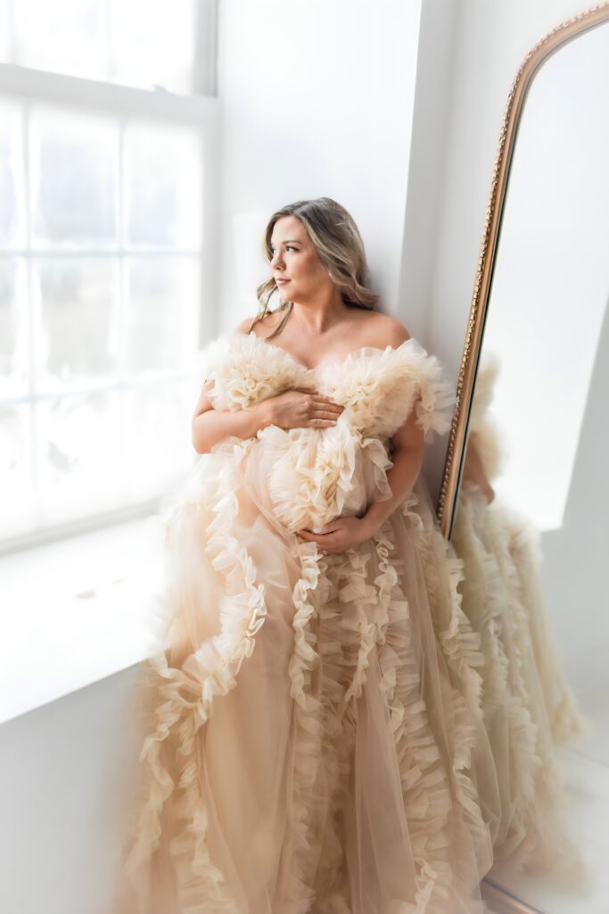 Studio maternity session featuring a glowing mother-to-be in a light, flowing gown, gently holding her baby bump.