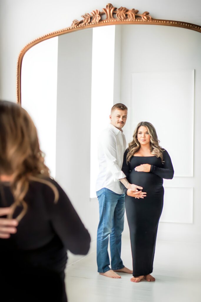 Maternity session capturing a peaceful moment of motherhood in a long black dress, surrounded by soft light with dad cradling bump.